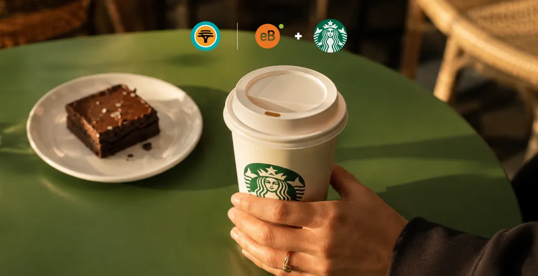 Hand holding a Starbucks takeaway coffee cup beside a chocolate brownie on a green café table, with eBucks and Starbucks partnership logos above in warm natural lighting.