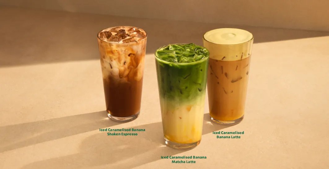 **Alt text:** Three iced Caramelised Banana Starbucks drinks in tall glasses on a warm beige surface: a shaken espresso, a matcha latte, and a latte, with soft golden light and shadows.